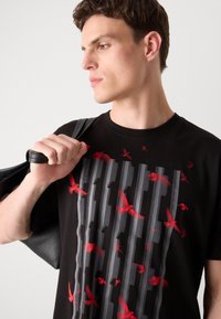 Schwarzes T-Shirt mit einer strukturierten Grafik aus grauen vertikalen Streifen und roten Vogelsilhouetten. Das Model hält eine schwarze Tasche über der Schulter.