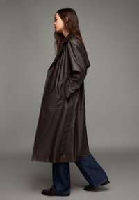 Brązowy płaszcz trencz z sztucznej skóry z dwurzędowymi guzikami i paskiem w talii, zestawiony z szerokimi dżinsami z niebieskiego denimu.