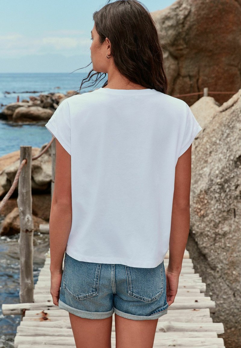 Frau mit langen dunklen Haaren, die ein weißes T-Shirt und Jeansshorts trägt, steht auf einem Holzsteg an einer felsigen Küste.