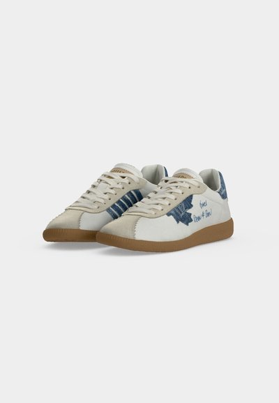 Baskets blanches et beige avec des patchs en denim bleu, texte style manuscrit sur les côtés, lacets beige et semelles en caoutchouc marron sur fond clair.