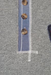 Grau Stoff mit strukturierter Oberfläche, ausgestattet mit einer blauen Blende, die vier braune Knöpfe und weiße Nähte aufweist. Keine zusätzlichen Details sichtbar.