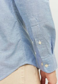 Camisa azul claro con un tejido suave y texturizado. Presenta un puño abotonado con dos botones y un dobladillo ligeramente redondeado.