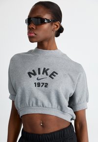 Pelēks īsais džemperis ar melnā krāsā izdrukātu "NIKE 1972", demonstrējot maigu tekstūru. Apvienots ar melnām saulesbrillēm un melnām biksēm.