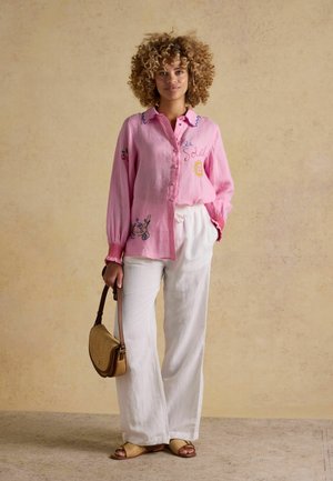 Frau mit lockigem Haar, die eine pinke bestickte Bluse, weiße weit geschnittene Hose, geflochtene Sandalen trägt und eine kleine braune Handtasche hält.