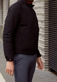 Zwarte gewatteerde jas met een hoge kraag, geribbelde schouderdetails en een rits aan de voorkant, gecombineerd met donkergrijze joggers.