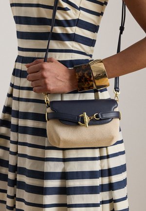 Femeie purtând o rochie în dungi navy și crem, ținând o geantă crossbody bej și navy cu închizătoare aurie, purtând brățări din scoică de țestoasă și aurii.