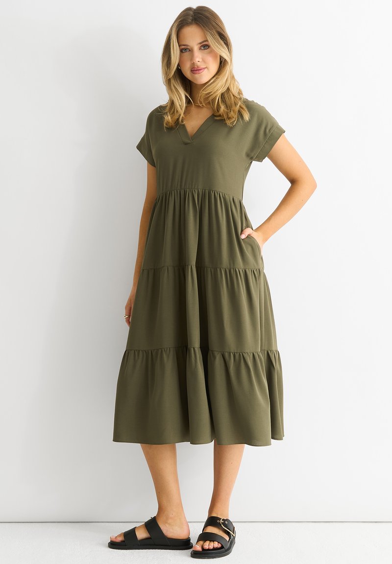 Olive green, short-sleeve dress with a V-neck, tiered ruffles, and side pockets; paired with black sandals, smooth fabric texture.