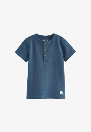 Camiseta de punto azul para niño pequeño, de manga corta, con cuello redondo, tres botones de madera en la tapeta y una pequeña etiqueta blanca de la marca cerca del dobladillo.