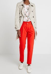 Cappotto trench beige sopra una maglietta a fantasia floreale, abbinato a pantaloni rossi brillanti e sneakers bianche. Bottoni piatti e vita definita sui pantaloni.
