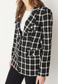 Mustassa ja valkoisessa ruudullisessa bleiserissä on kultaiset napit, rakenteelliset kaulukset, kaksi etutaskua ja teksturoitu kangas. Symmetriallinen kuvio koko vaatteessa.