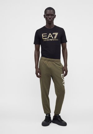 Homme portant un t-shirt noir EA7 Emporio Armani et un pantalon de survêtement vert olive avec des baskets noires, debout devant un fond blanc uni.