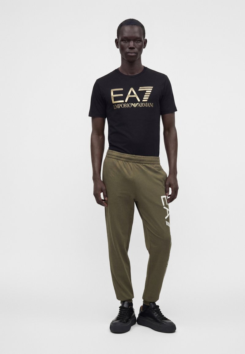 Άνδρας που φοράει μαύρο t-shirt EA7 Emporio Armani και ελιά πράσινο παντελόνι φόρμας με μαύρα αθλητικά, στέκεται μπροστά σε λευκό μονόχρωμο φόντο.