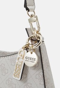 Helle graue, strukturierte Handtasche mit schwarzem Akzent, ausgestattet mit goldfarbenen Beschlägen, einem Logo-Anhänger und einem runden Charm, der mit "GUESS EST.1981." beschriftet ist.