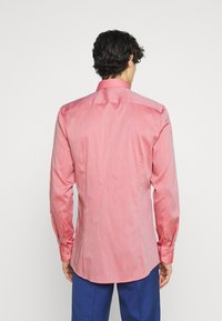 Uomo con capelli ricci scuri che indossa una camicia a maniche lunghe rosa e pantaloni blu, mostrato di spalle su uno sfondo bianco.