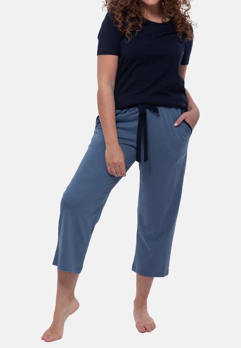 Kvinna iklädd en marinblå kortärmad skjorta och blå capribyxor med dragsko, som står barfota med en hand i fickan och ett ben något lyft.