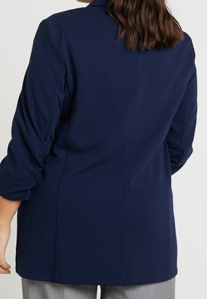 Personne aux cheveux bruns mi‐longs portant un blazer bleu marine et un pantalon gris clair, vue de dos sur fond blanc.