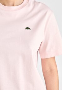 Svetlo roza bombažna majica s kratkimi rokavi, z majhnim zelenim vezanjem logotipa krokodila na levem prsnem delu.