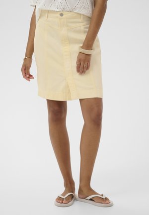 Kvinde iført en bleg gul knælang nederdel, hvid bluse med hulmønster, beige klipklappere og guld- og beige armbånd på hænderne.