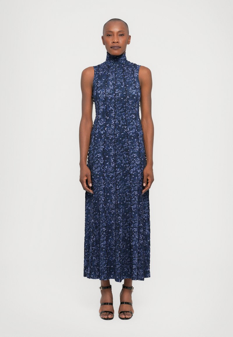 Rochie fără mâneci, cu imprimeu floral într-o nuanță de albastru marin, gât înalt, design pliat și fustă până la gleznă. Asortată cu sandale negre cu bretele.