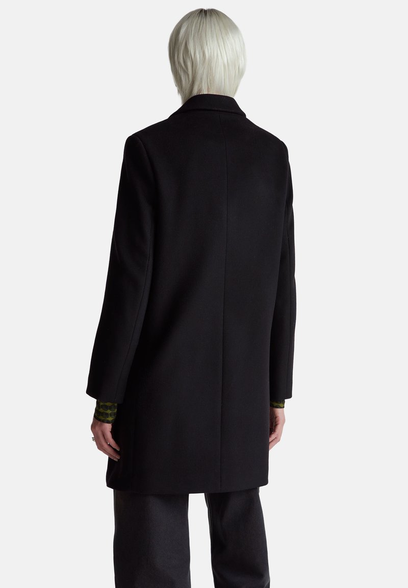 Manteau noir avec une coupe ajustée, manches longues et une fente unique à l'arrière. Le tissu semble lisse avec un design structuré.