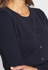 Kvinna som bär en marinblå knappad kofta med trekvartsärmar, med armarna i kors och en naturlig hudton synlig på hals och hand.