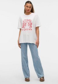 Camiseta blanca de algodón con un diseño gráfico rojo que presenta langostas y el texto "LA DOLCE VITA", combinada con unos jeans de pierna ancha en azul claro y zapatos marrones.