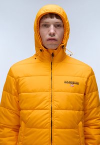 Gelb gefütterte Pufferjacke mit gestepptem Muster, Frontreißverschluss und Logo auf der Brust. Verfügt über Zugbänder in der Kapuze zur Anpassung.