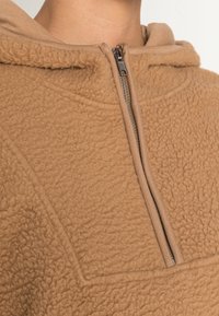 Pullover in pile marrone con zip a metà davanti. Presenta una superficie testurizzata e un bordo cappuccio liscio. Design semplice e pratico.