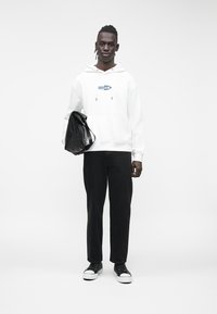 Camisola com capuz branca com logo azul "Guess Jeans", calças pretas e mochila preta. Ténis pretos com detalhes em branco. Design simples e moderno.