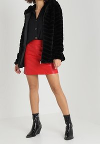 Černá umělá kožešinová bunda s kapucí a žebrovanou texturou, nošená přes krátkou červenou sukni. Černé lesklé kotníkové boty s klínovým podpatkem dokončují vzhled.