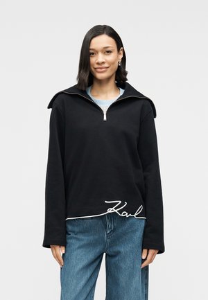 Svart genser med høy krage, halvlange glidelås og hvit kursiv logo ved kanten. Har lange ermer og en avslappet passform.