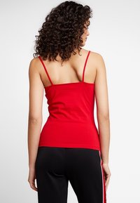 Topino rosso aderente con spalline sottili, realizzato in un tessuto liscio, caratterizzato da un design semplice e una schiena senza cuciture. Indossato con pantaloni neri.