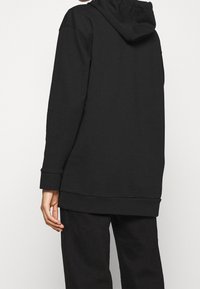 Černá mikina s kapucí volnějšího střihu, vyrobená z měkkého materiálu. Má kapuci, dlouhé rukávy a žebrované manžety a lem.