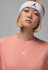 Lys rosa t-skjorte med rund hals og teksturert stoff, med en hvit Jordan-logo på brystet. Hvit hårbånd med svart logo.