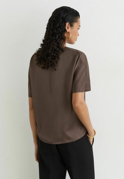 Woman with curly dark hair wearing gold hoop earrings and brown short-sleeve top, standing with one hand in pocket against white wall.