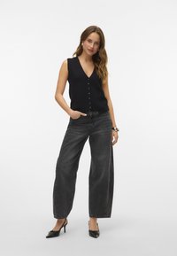 Top in maglia nera senza maniche con scollatura a V e chiusura con bottoni, abbinato a jeans neri a vita alta e gamba larga e tacchi neri.