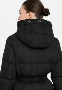Giacca a vento nera con cappuccio, caratterizzata da un design trapuntato, una vita elastica e un dettaglio colletto di paillettes. Texture liscia e opaca.