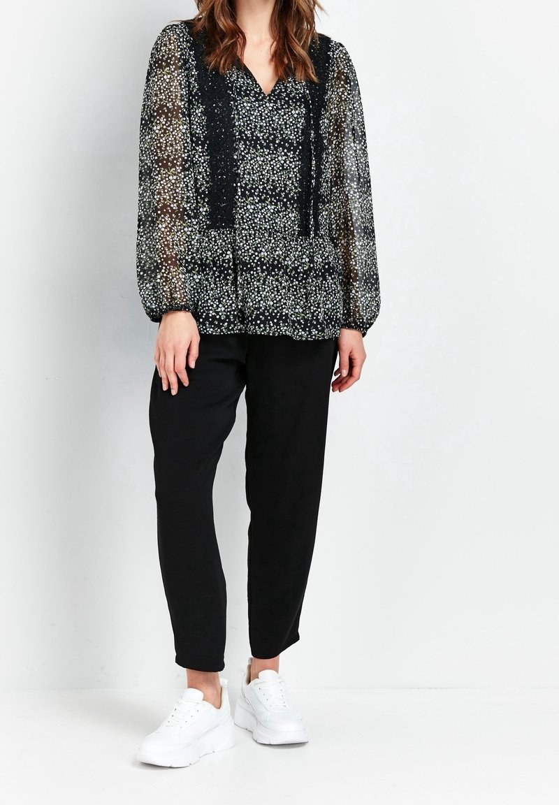 Transparante zwarte blouse met witte en groene bloemmotieven, met een V-hals en geplooide mouwen, gecombineerd met nauwsluitende zwarte broek en witte sneakers.