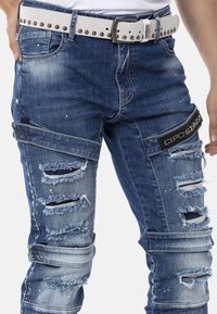 Blaue Jeans aus Denim weisen beschädigte Patches, ausgefranste Kanten und einen weißen Nietengürtel auf. Ein schwarzes Etikett an der Seite fügt ein dekoratives Detail hinzu.