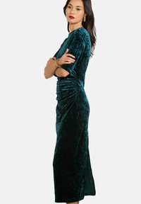 Žena s dlhými tmavými vlasmi, oblečená v tehlovej sametovej midi šatovej s riaseným pásom, stojí so založenými rukami na neutrálnom pozadí.