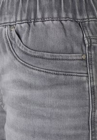 Harmaat farkut, joissa on ommeltu tasku, metallinen neppari ja teksturoitu kangaspinta. Vyötärönauha on joustava mukavuutta varten.