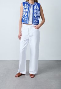 Blå broderet cropped vest med hvide blomsterprint, kombineret med løse hvide bukser og brune sandaler med stropper på en grå baggrund.