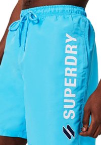 Bañador azul claro con cintura elástica y cordón ajustable. "SUPERDRY" está impreso en blanco verticalmente en el lateral, con un acento negro.
