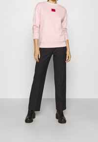 Sudadera rosa con un parche de logo rojo, combinada con pantalones rectos negros y botas de tobillo negras. Textura de tela suave, ajuste casual.