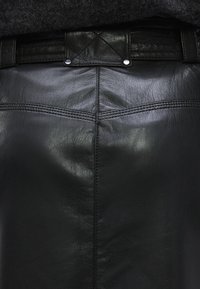 Mustast nahast pliiatsseelik, millel on sile tekstuur, vertikaalsed õmblused ning taljeosa, mis on kaunistatud tikandiga ja metallist trukkidega.