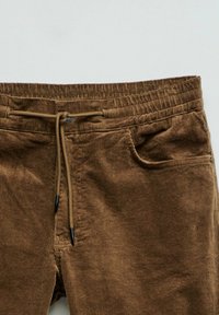 Smeđe kord pantalone s teksturiranom površinom, elastičnim strukom i zatvaranjem na vezicu. Ima bočne džepove za dodatnu funkcionalnost.