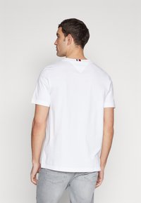 T-shirt bianco in cotone con maniche corte, caratterizzato da un dettaglio a yoke triangolare sulla schiena e una piccola striscia di accento rossa, bianca e blu.