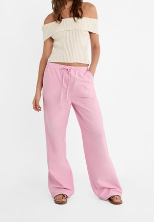 Woman wearing cream off-shoulder ribbed knit top, loose pink drawstring pants, and brown flat sandals, standing with one hand in pocket.