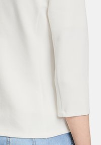 Biała bluzka o teksturze z rękawami ¾, z okrągłym dekoltem i prostym dołem. Materiał ma subtelny wzór, co nadaje jej strukturalny wygląd.