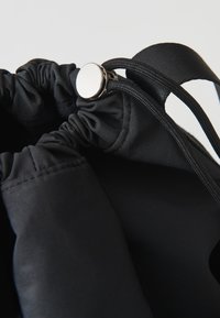 Sacca nera con coulisse, dal fondo liscio e opaco, con apertura arricciata, presenta un fermo metallico rotondo e spallacci a contrasto.